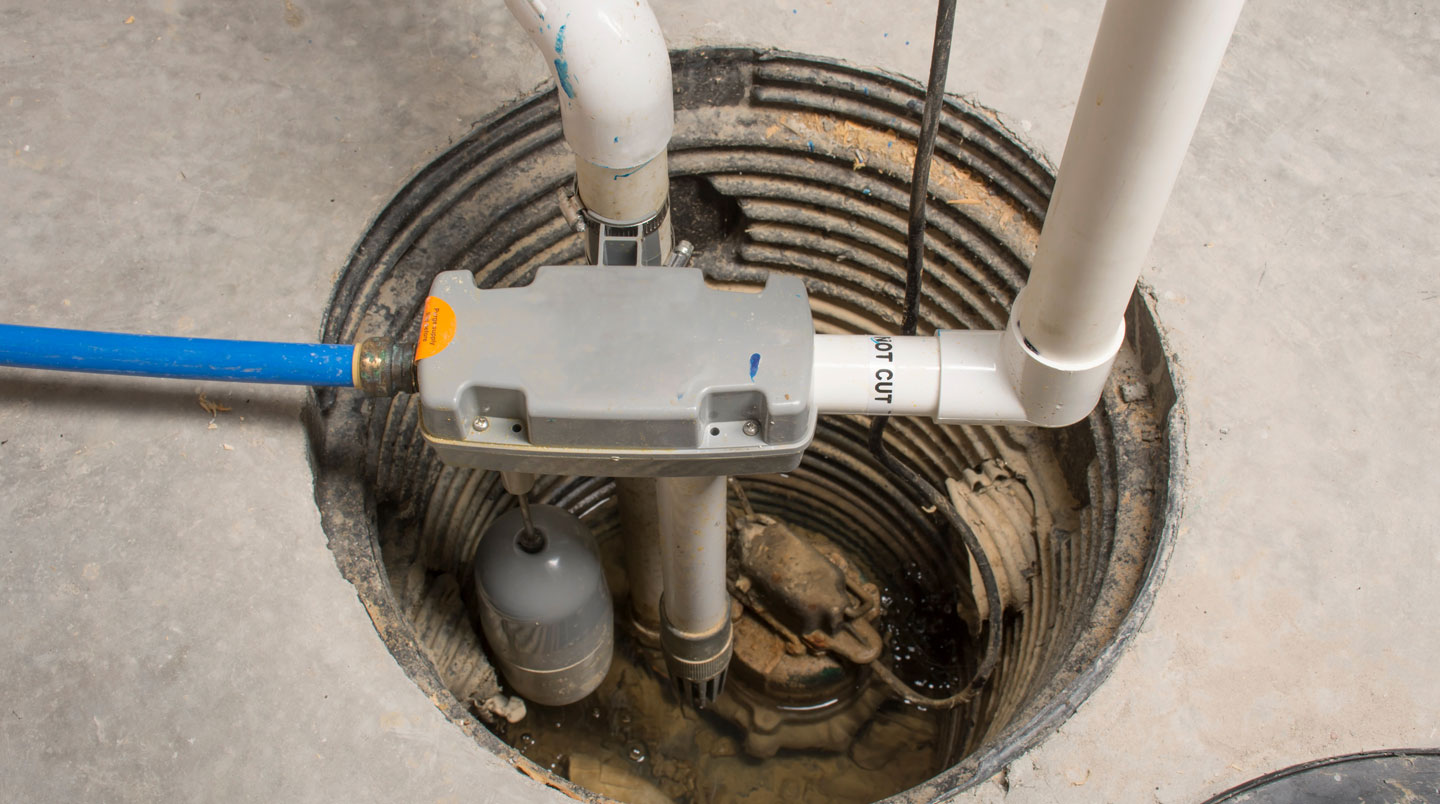 Pompe de puisard submersible noire installée dans un bassin circulaire en béton au sol d'un sous-sol résidentiel, avec tuyau d'évacuation PVC blanc montant verticalement vers le plafond